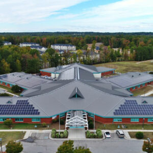 An aerial photo shows a public school in Richmond, Virginia, with solar panels on several sections of the roof. Houses and woods are visible in the distance.