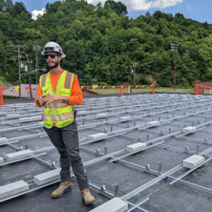 A male worker in a neon construction vest, white hard hat, work boots and sunglasses stands on a roof being prepared for solar panel installation.