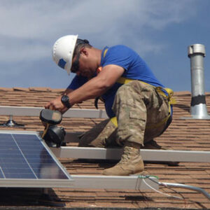 A man in a white hard hat, blue t-shirt and camouflage pants installs a solar panel on a rooftop.
