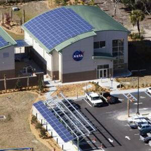 Solar panels cover the rounded roof of a NASA building. Another solar array shades the parking lot. Palm trees are visible in the background.