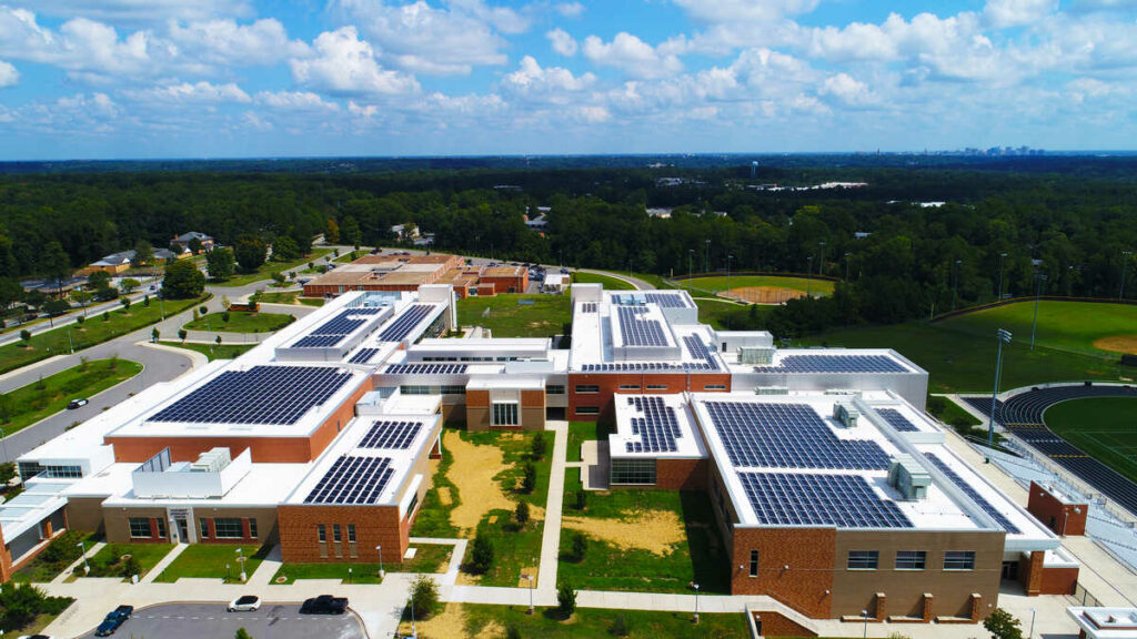 A wide aerial photo of Huguenot High School shows solar panels on roofs across the facility with sports fields in the distance.