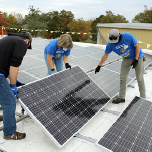 Three volunteers in blue t-shirts install a solar panel on the roof of Eastern Mennonite School.