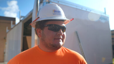 Apprentice solar technician wearing a white hard hat and an orange shirt.