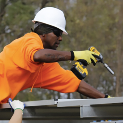 Man wearing a white hard hat and an orange t-shirt holds a power drill while installing solar panels.