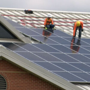 Two men in orange shirts and hard hats install solar panels on the roof of Linwood Holton Elementary School.