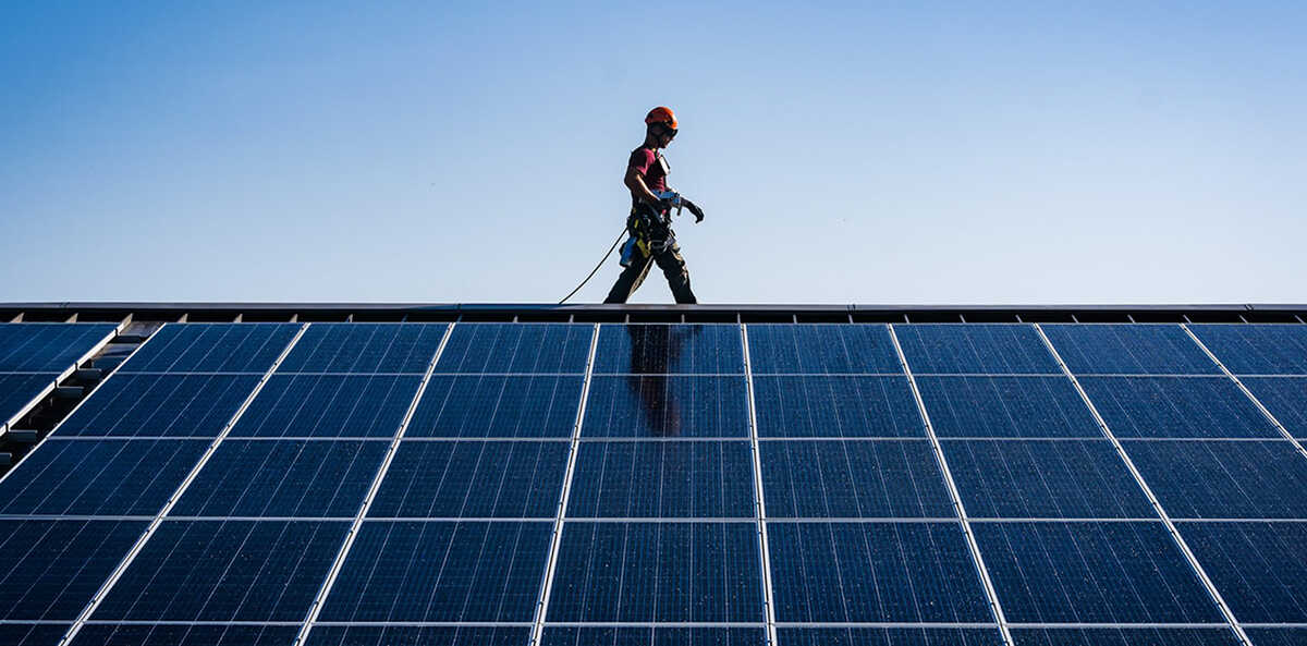 A man wearing a hard hat walks along the top of a solar panel against a backdrop of blue sky.