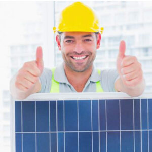 A man in a yellow hard hat makes two thumbs up signs next to a solar panel.