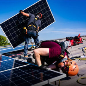 A photo of two men in construction gear installing solar panels on the roof of a building. One man is carrying a solar panel while the other is kneeling down to secure a panel to the roof.