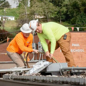 Two solar apprentices work together to install solar power equipment on the roof of a public school building.