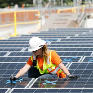 A female worker in a yellow vest and white hard hat uses blue tape to mark solar panels on the roof of Huguenot High School in Richmond, Virginia.