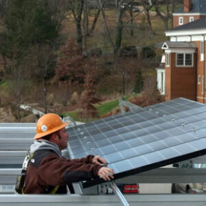 A worker in an orange hard hat and winter jacket installs a solar panel on the roof of a parking garage. Brick college buildings can be seen in the background.