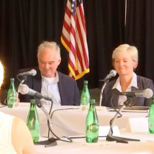 U.S. Sen. Tim Kaine and U.S. Secretary of Energy Jennifer Granholm sit at a banquet table in Troutville, Virginia. An American flag can be seen in the background and green glass water bottles sit on the table.
