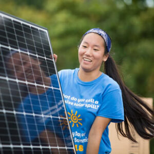 A smiling Asian American female college student in a blue t-shirt carries one end of a solar panel with a partner who is out of the frame. Her reflection can be seen in the surface of the solar panel.