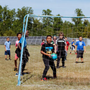 A group of young boys plays soccer in a field with solar panels in the background. The goalie smiles at the camera through the netting.