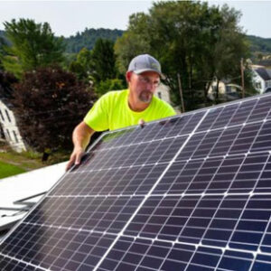 A man in a green t-shirt and gray baseball cap is installing a solar panel on the roof of a building in West Virginia.