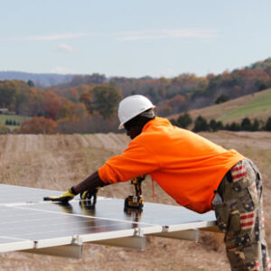 An African American worker in an orange shirt, camouflage pants and white hard hat installs solar panels in a farm field.
