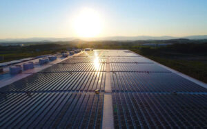 The setting sun shines on a large array of roof mounted solar panels on a warehouse.